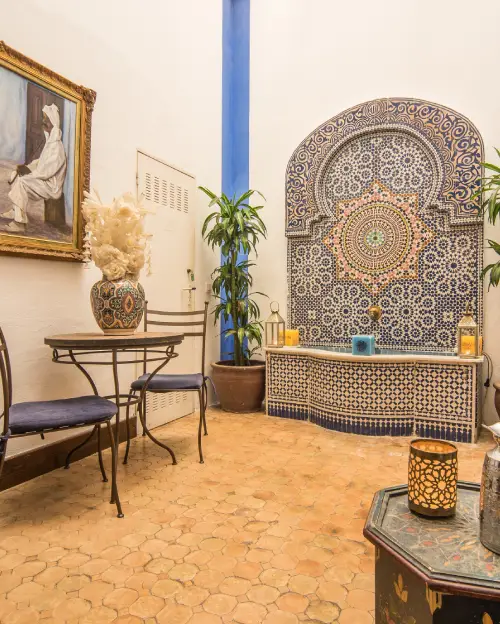 Patio con fuente de mosaicos, plantas y mesa con sillas de hierro forjado.