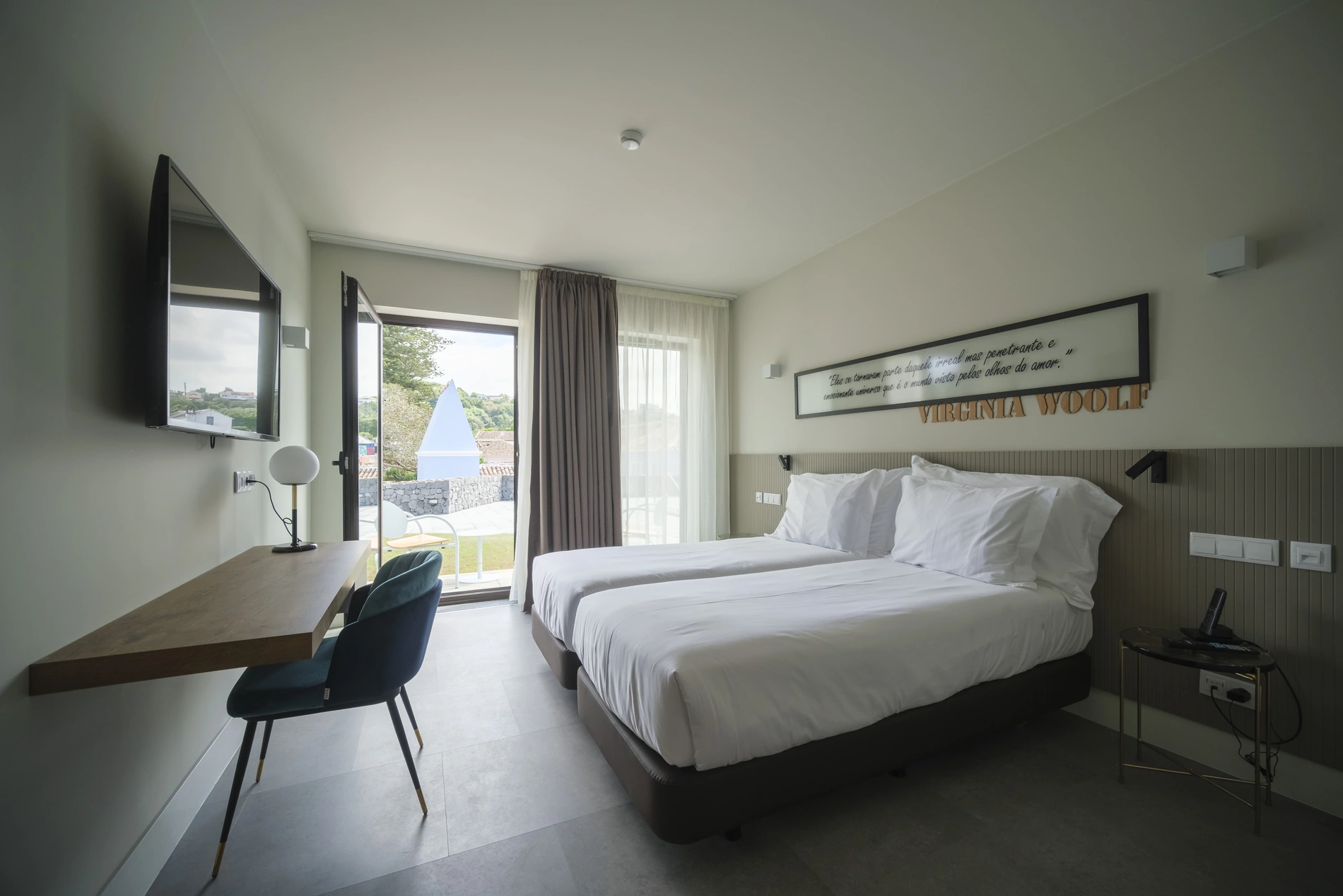 Quarto de hotel com cama de casal, mesa, TV e vista para a &aacute;rea externa.