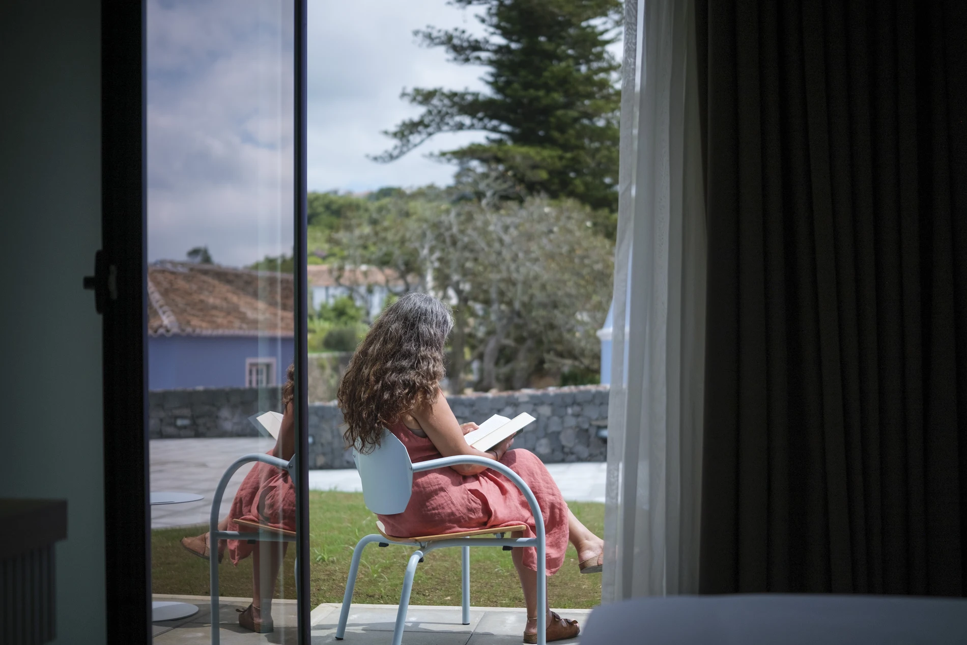 Mulher sentada em cadeira ao ar livre, lendo livro sob o sol.