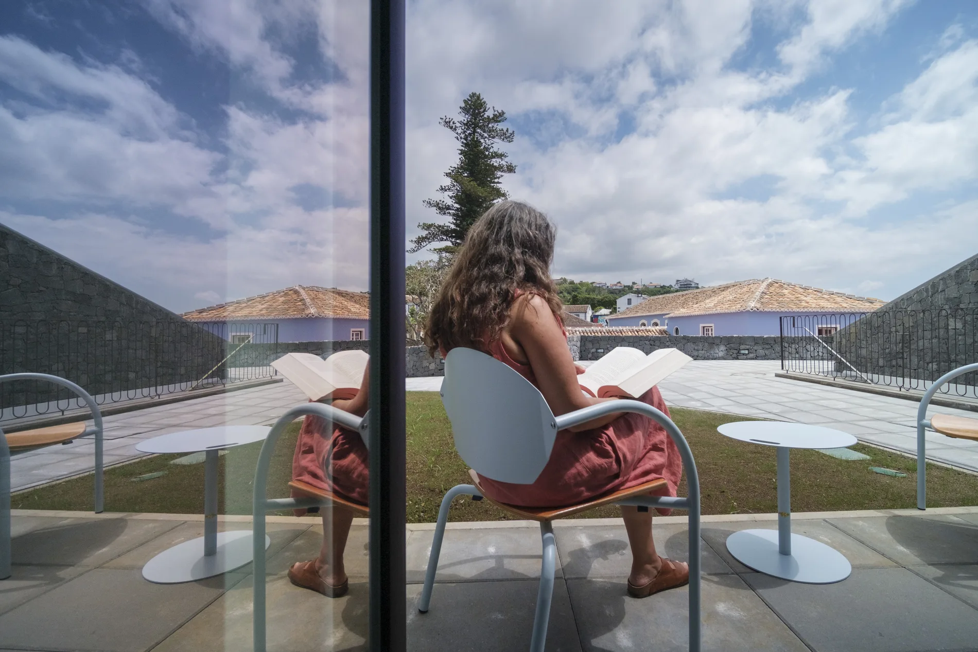Mulher lendo em uma cadeira com reflexo em vidro ao ar livre.