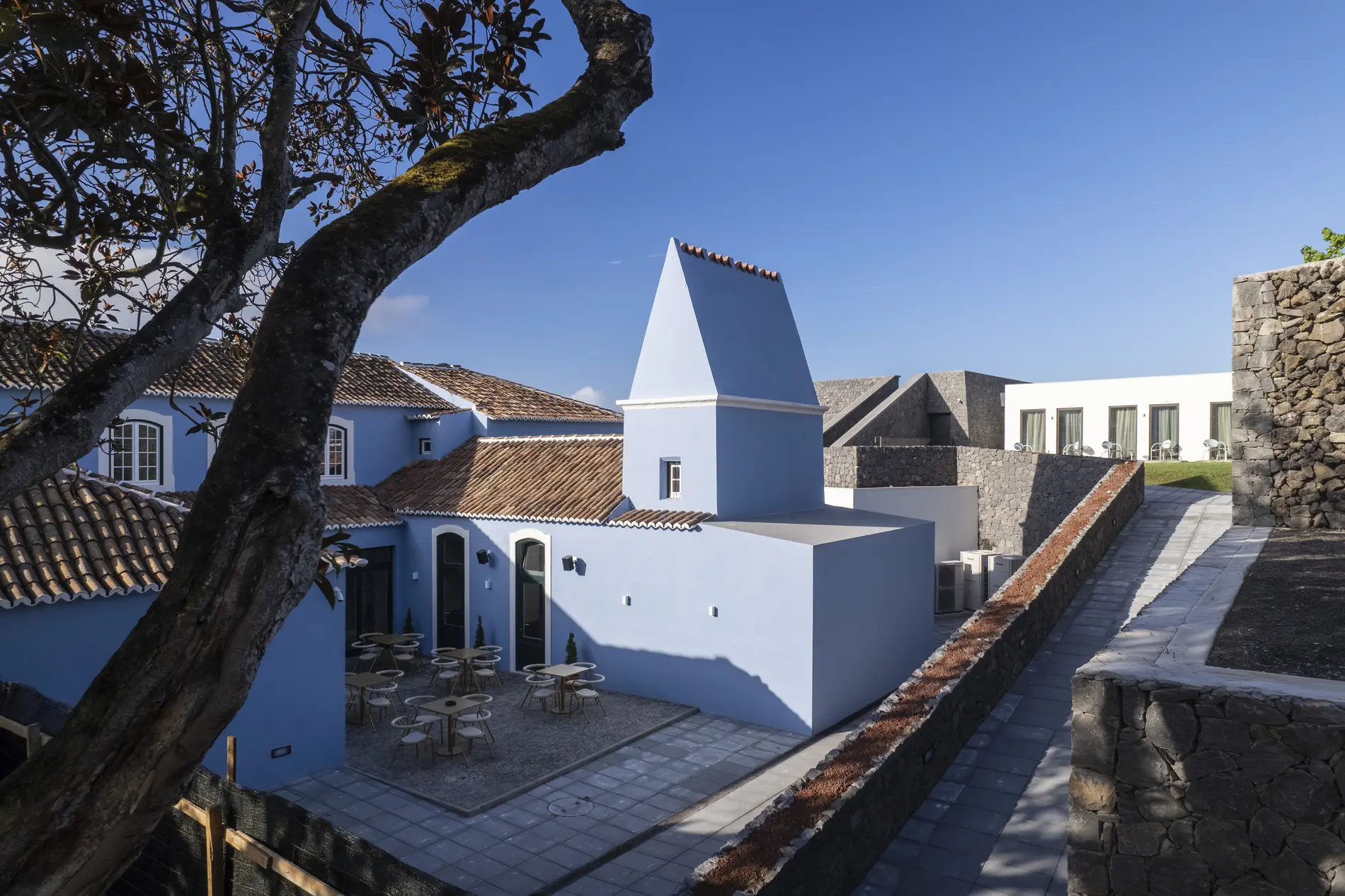 Edif&iacute;cios azul e pedra com p&aacute;tio e &aacute;rvores sob c&eacute;u azul.
