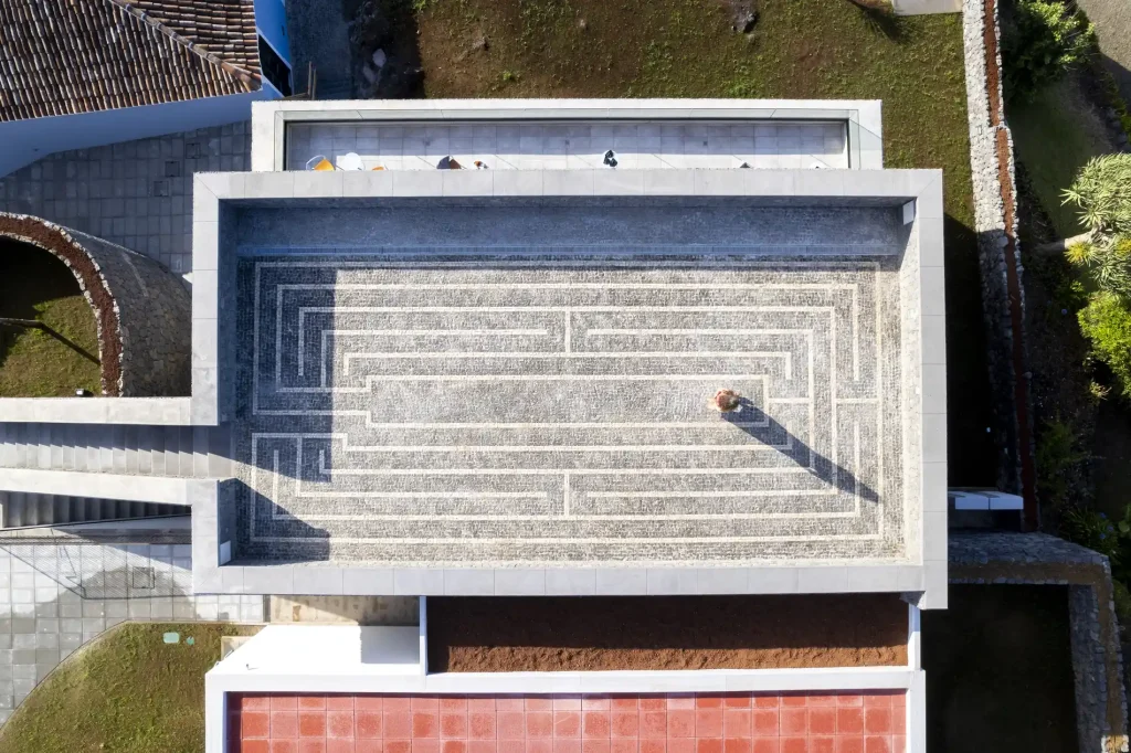 Vista a&eacute;rea de um p&aacute;tio com mosaico, sombra central e vegeta&ccedil;&atilde;o ao redor.