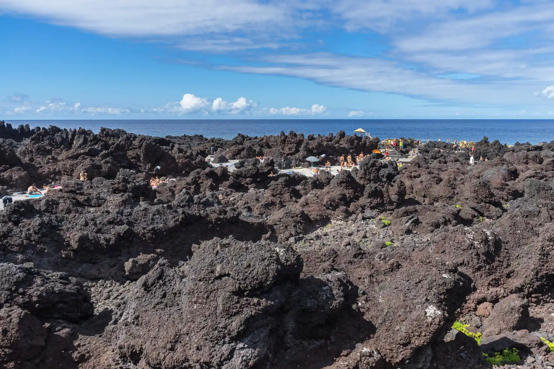Forma&ccedil;&otilde;es de lava e pessoas sob sol com c&eacute;u azul e nuvens ao fundo.