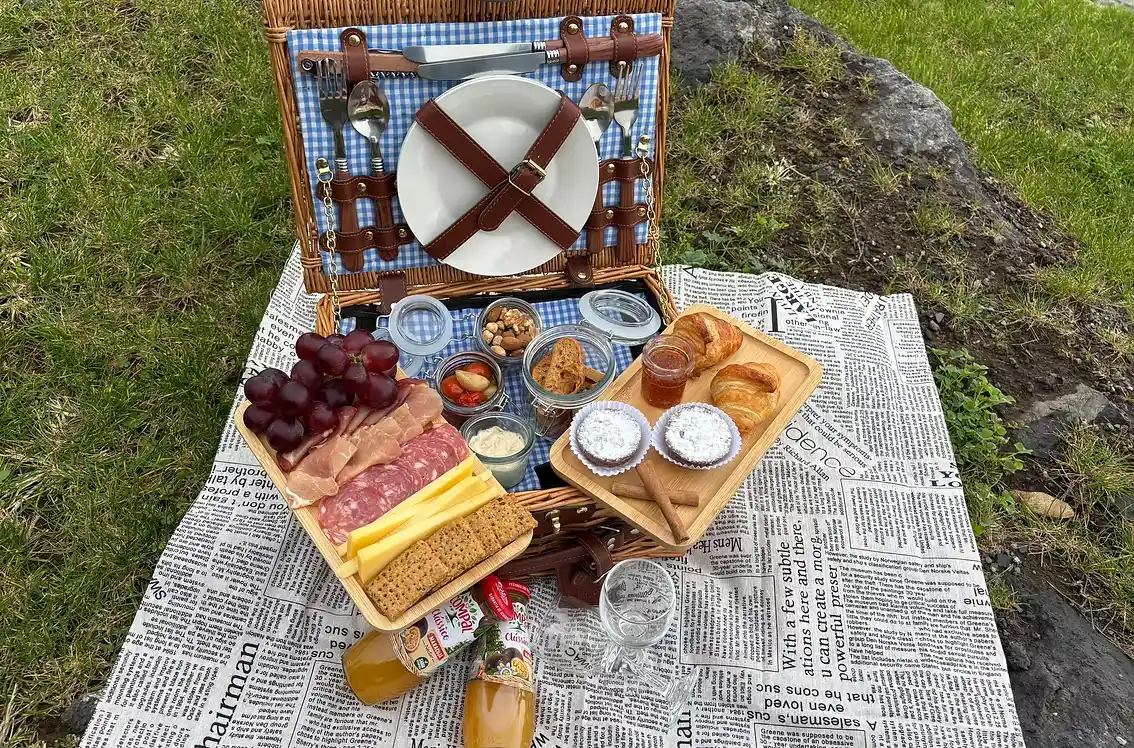 Cesta de piquenique com frios, p&atilde;es, frutas e bebidas sobre jornal na grama.
