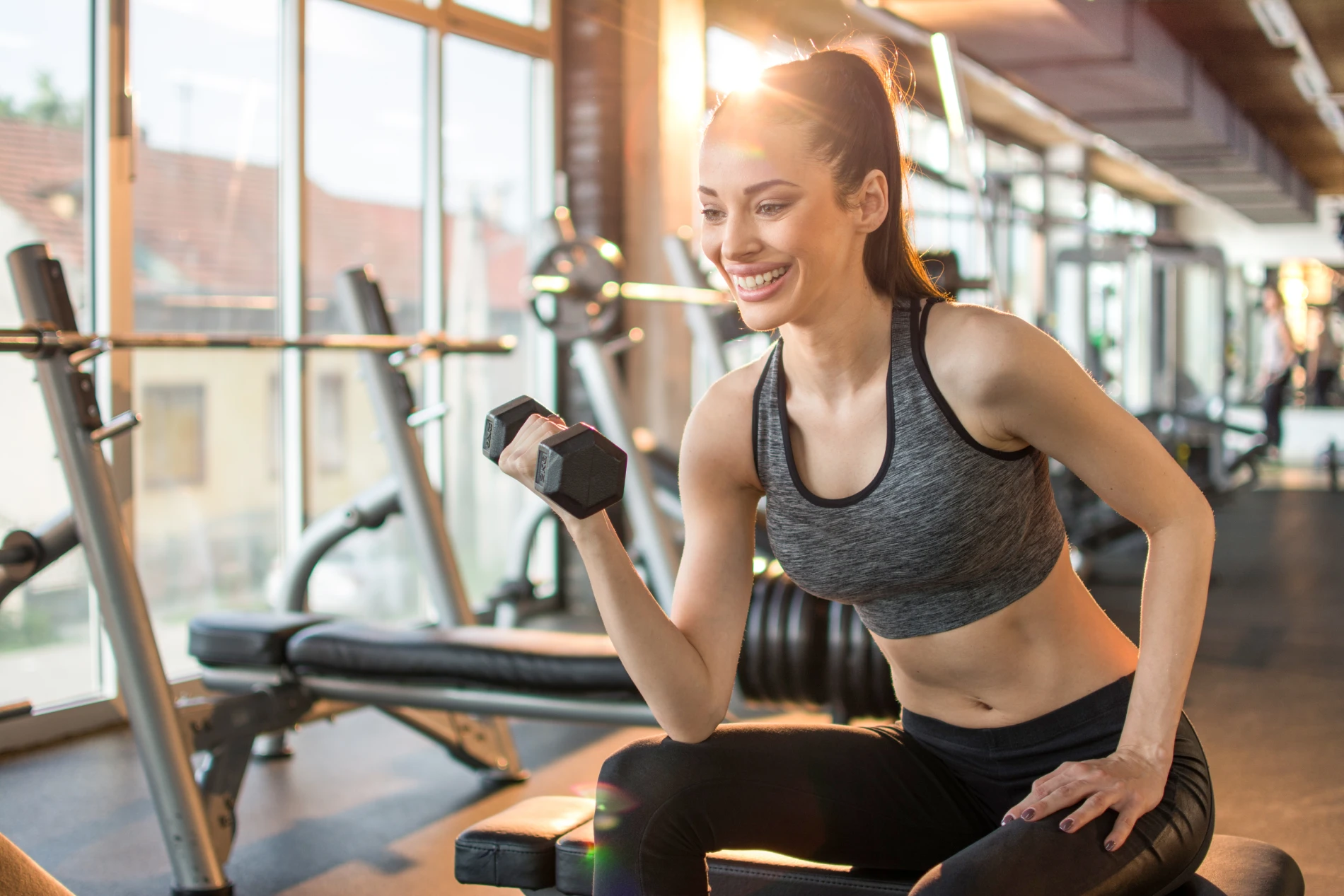 Mulher sorrindo levantando haltere em uma academia iluminada pelo sol.