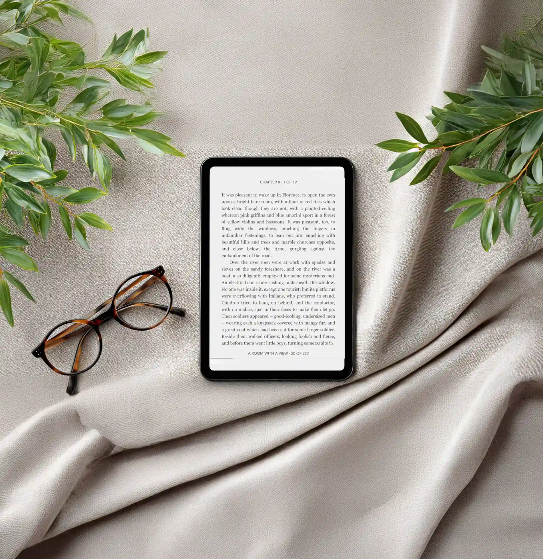 &Oacute;culos e tablet com texto sobre tecido claro, cercados por folhas verdes.