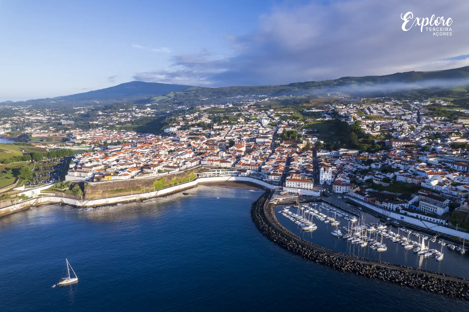 Vista a&eacute;rea de uma cidade costeira com marina e montanhas ao fundo.