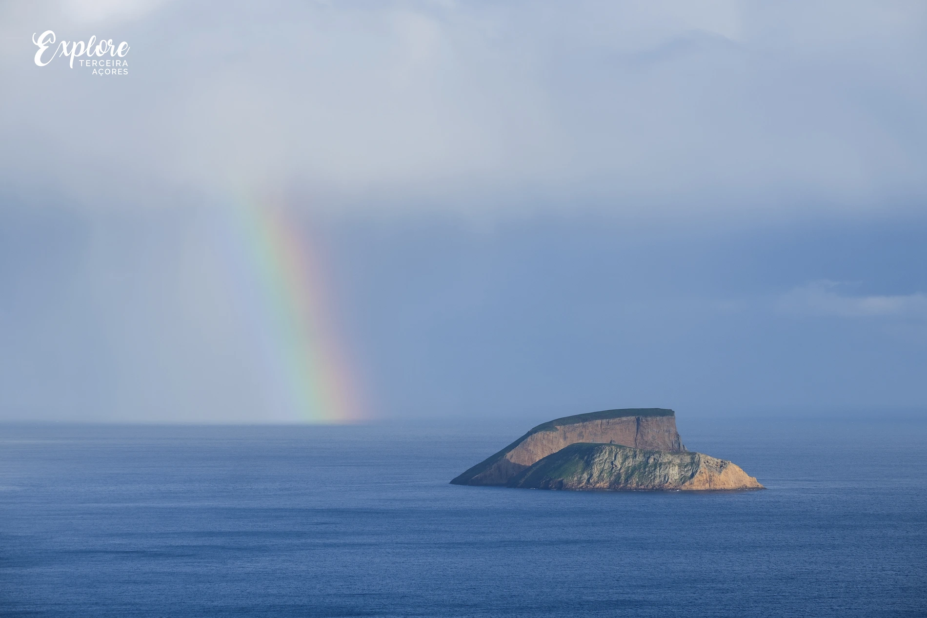 Arco-&iacute;ris sobre o mar com uma ilha rochosa ao fundo.