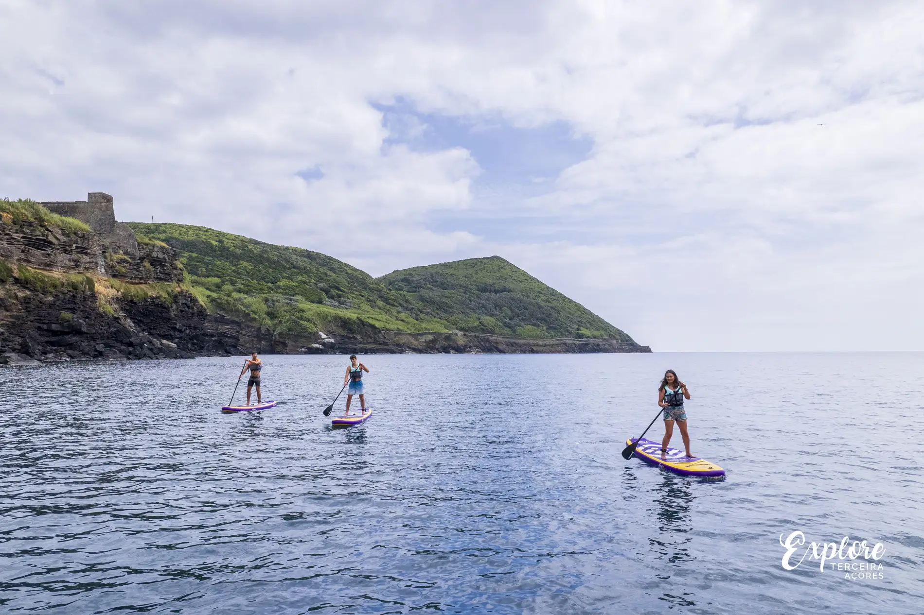Tr&ecirc;s pessoas praticam stand-up paddle no mar com colina ao fundo.