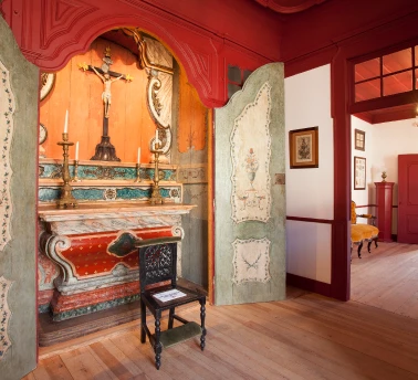 Capela antiga com altar ornamentado e cadeira diante de porta entreaberta.