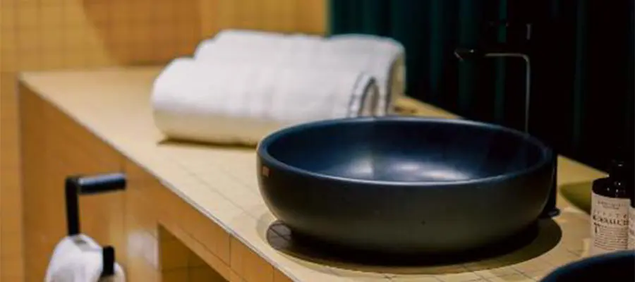 Lavabo negro en ba&ntilde;o con toallas blancas y jab&oacute;n sobre encimera amarilla.