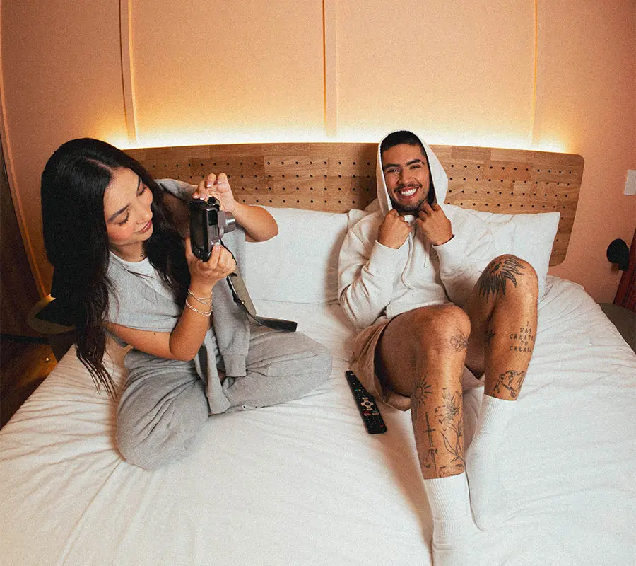 Persona sonriente en una cama mientras otra toma una foto, ambos relajados.