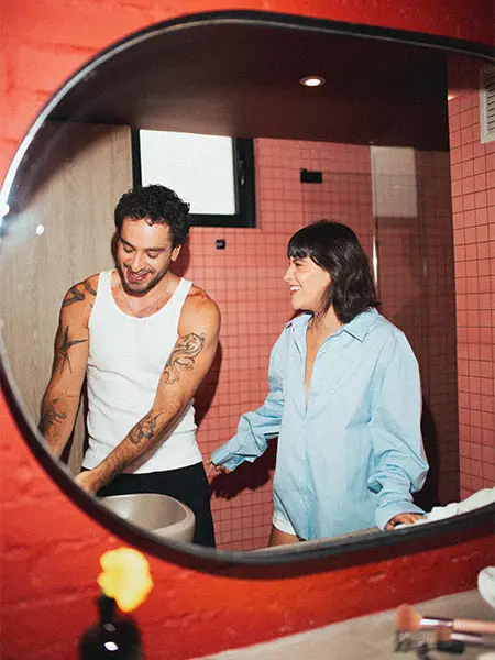 Pareja sonriendo frente al espejo en un ba&ntilde;o con azulejos rosas.