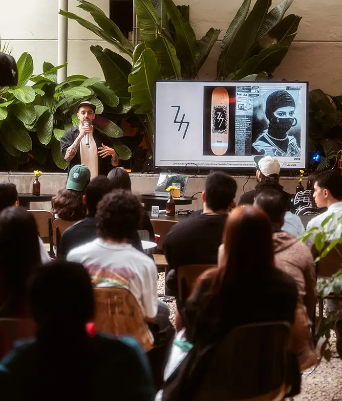 Persona dando una presentaci&oacute;n en exterior con p&uacute;blico, pantalla y plantas al fondo.