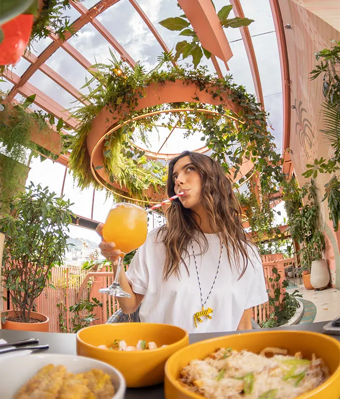 Mujer con bebida en terraza decorada con plantas y platos de comida en primer plano.