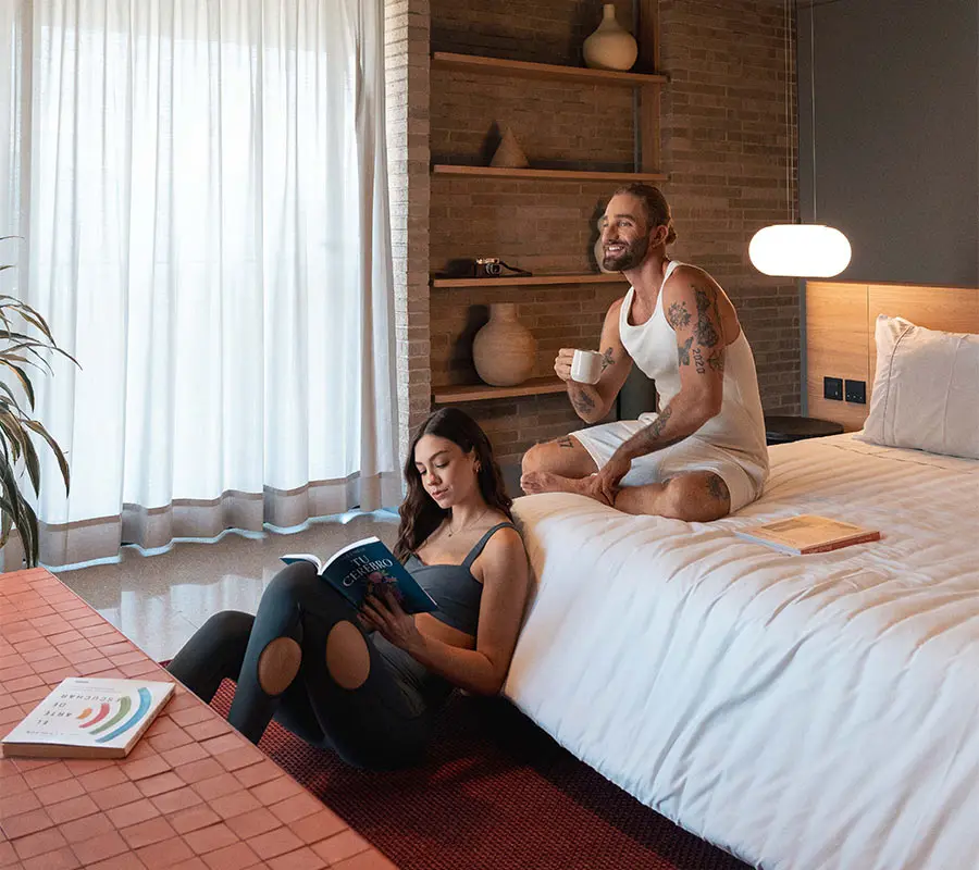Mujer leyendo en el suelo y hombre tomando caf&eacute; en la cama en un dormitorio acogedor.