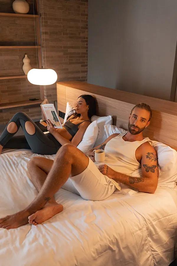 Pareja relaj&aacute;ndose en la cama; ella lee, &eacute;l toma caf&eacute;.