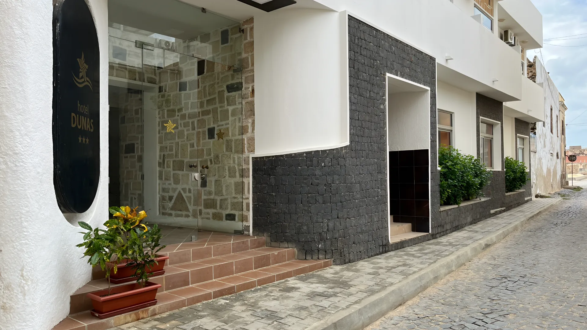 Entrada de hotel com fachada de pedra, plantas em vasos e rua de paralelep&iacute;pedos.