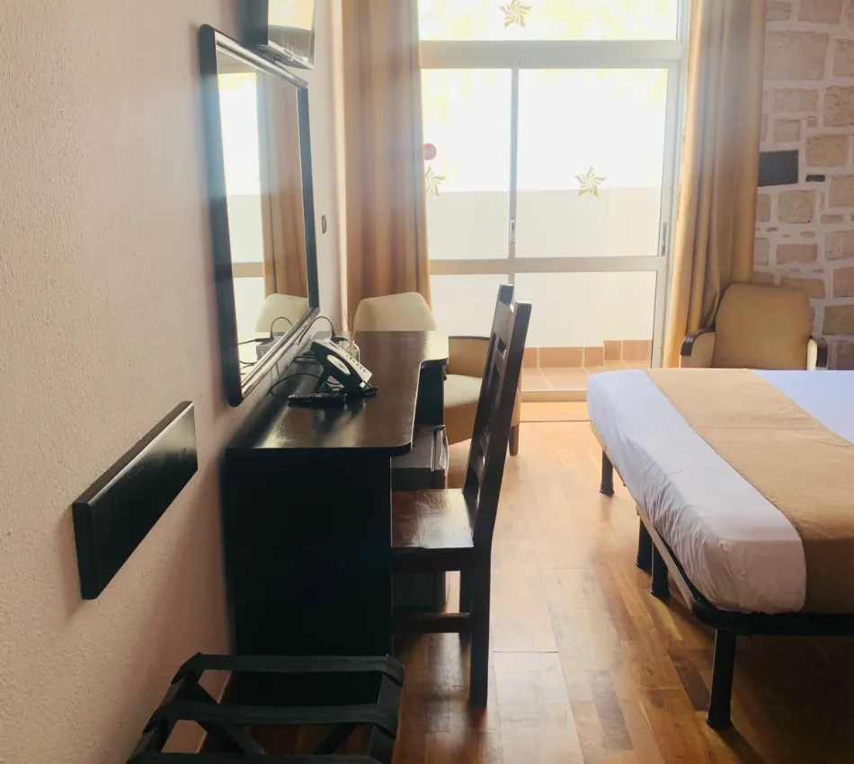 Quarto de hotel com cama, mesa, telefone, espelho e janela com cortinas.