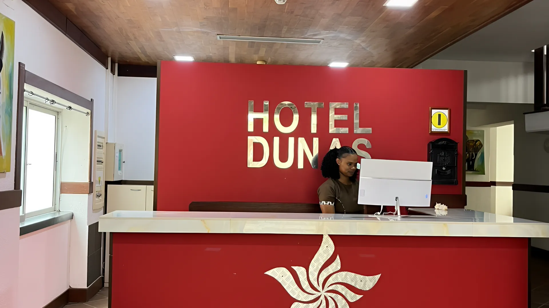 Recep&ccedil;&atilde;o do Hotel Dunas com atendente ao computador, fundo vermelho e logotipo na parede.