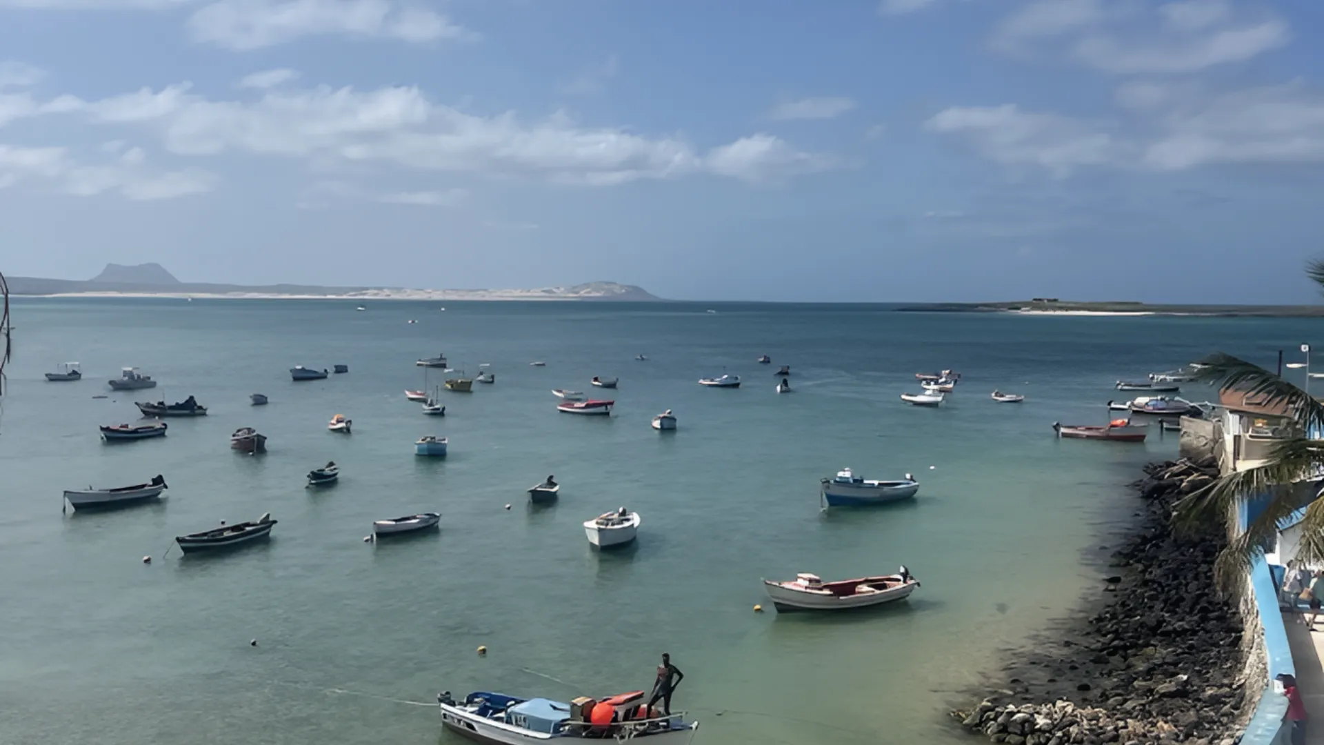 Barcos ancorados em uma ba&iacute;a tranquila, com c&eacute;u azul e montanha ao fundo.