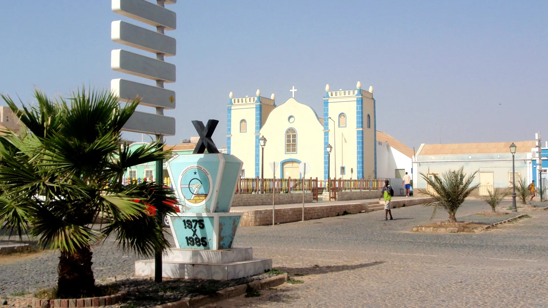 Edif&iacute;cio hist&oacute;rico de cor azul e branca com palmeiras e monumento em primeiro plano.