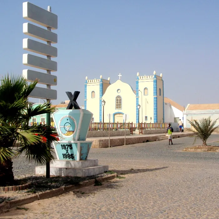 Monumento e igreja hist&oacute;rica em vila costeira, com c&eacute;u limpo e palmeiras ao redor.