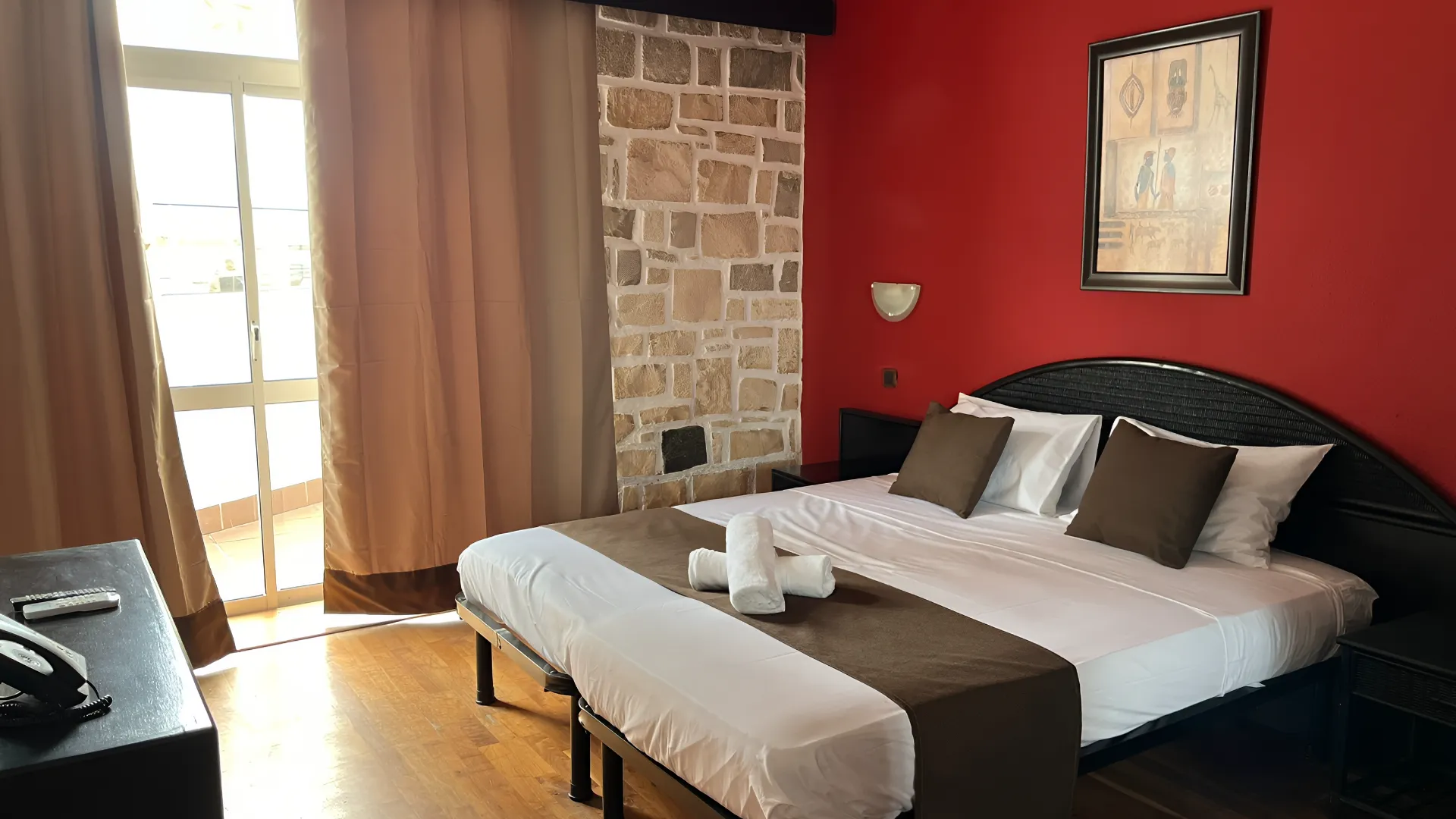 Quarto de hotel com cama de casal, paredes vermelha e de pedra, e janela com cortinas claras.