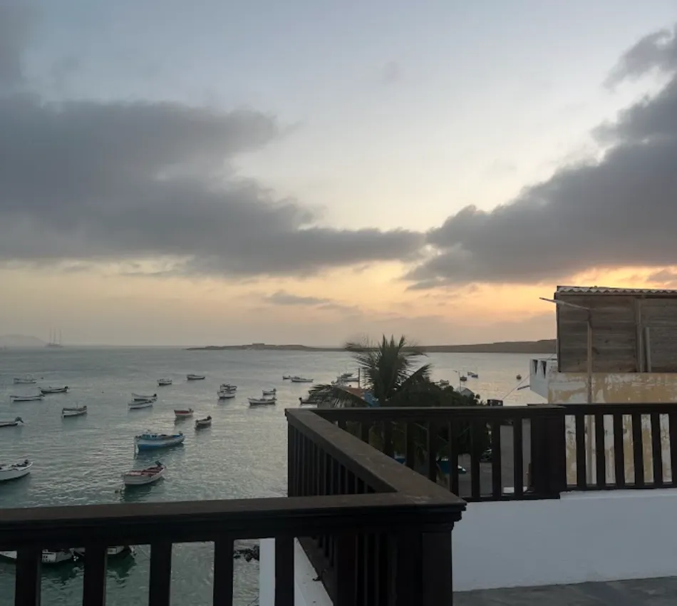 Vista de um p&ocirc;r do sol sobre o mar com barcos e c&eacute;u nublado, visto de uma varanda.