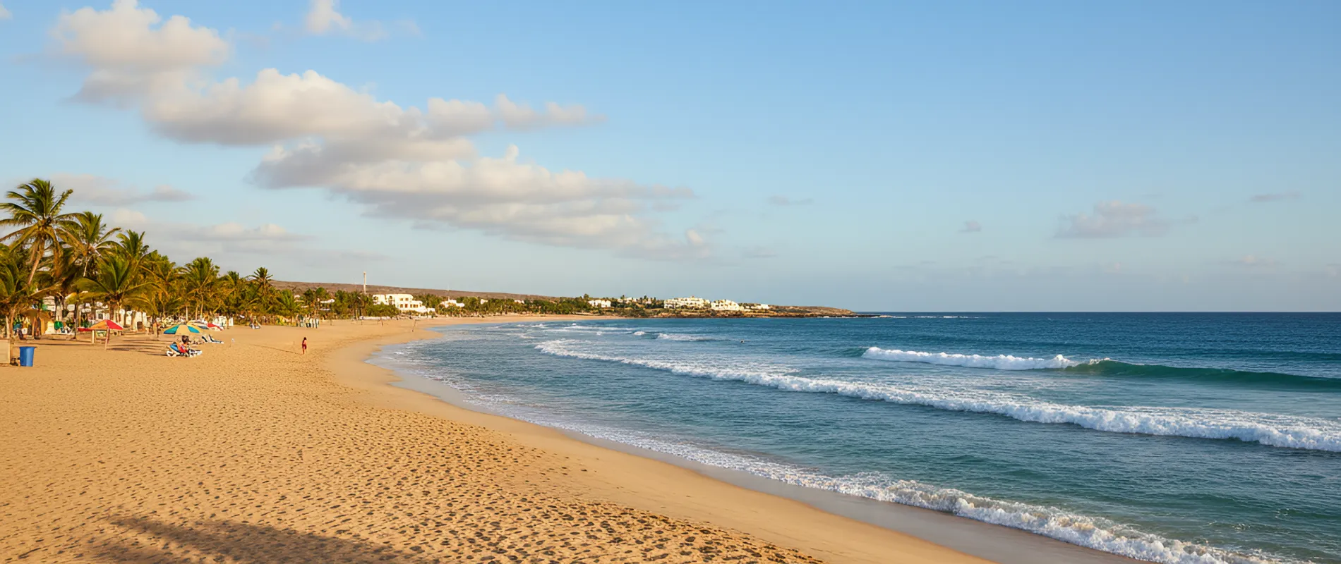 Praia com areia dourada, palmeiras e mar azul sob c&eacute;u claro e poucas nuvens.