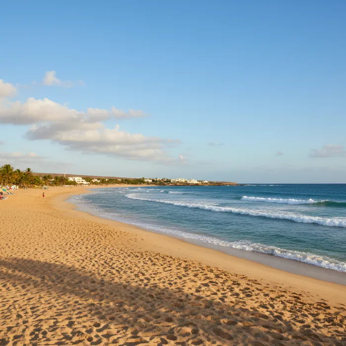 Praia com areia dourada, mar azul e c&eacute;u claro com algumas nuvens.