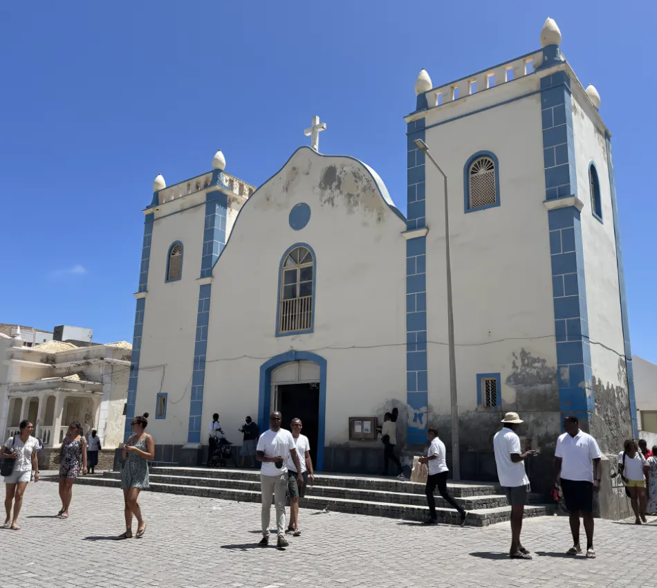 Igreja com fachada desgastada em pra&ccedil;a de pedra, pessoas ao redor, c&eacute;u azul.