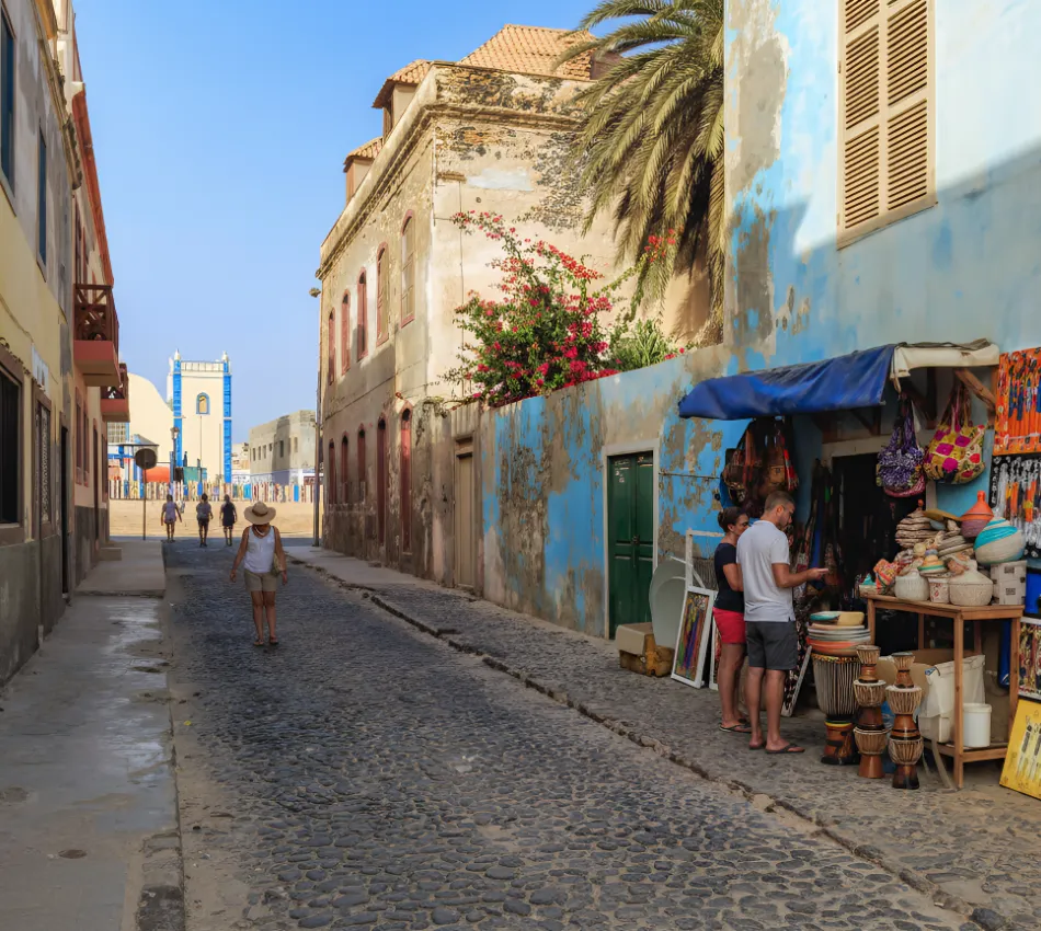 Rua de paralelep&iacute;pedos com pr&eacute;dio azul, pessoas caminhando e vendinha com artesanatos coloridos.