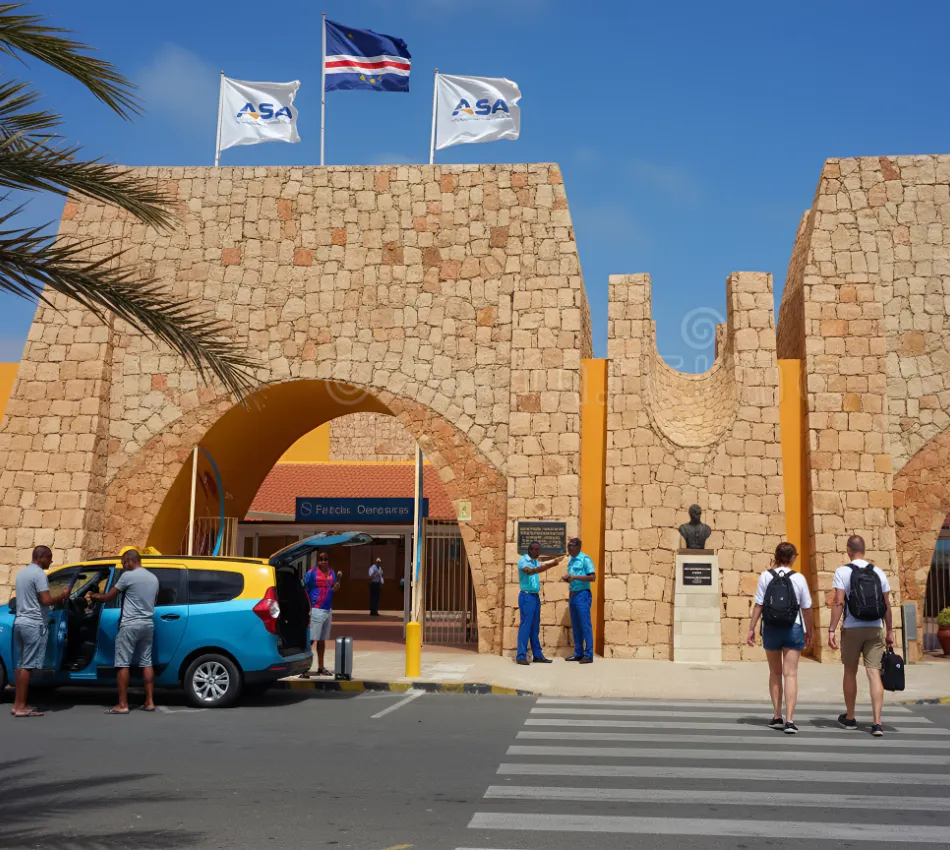 Entrada de aeroporto com pessoas e t&aacute;xi; bandeiras ao vento sobre estrutura de pedra.