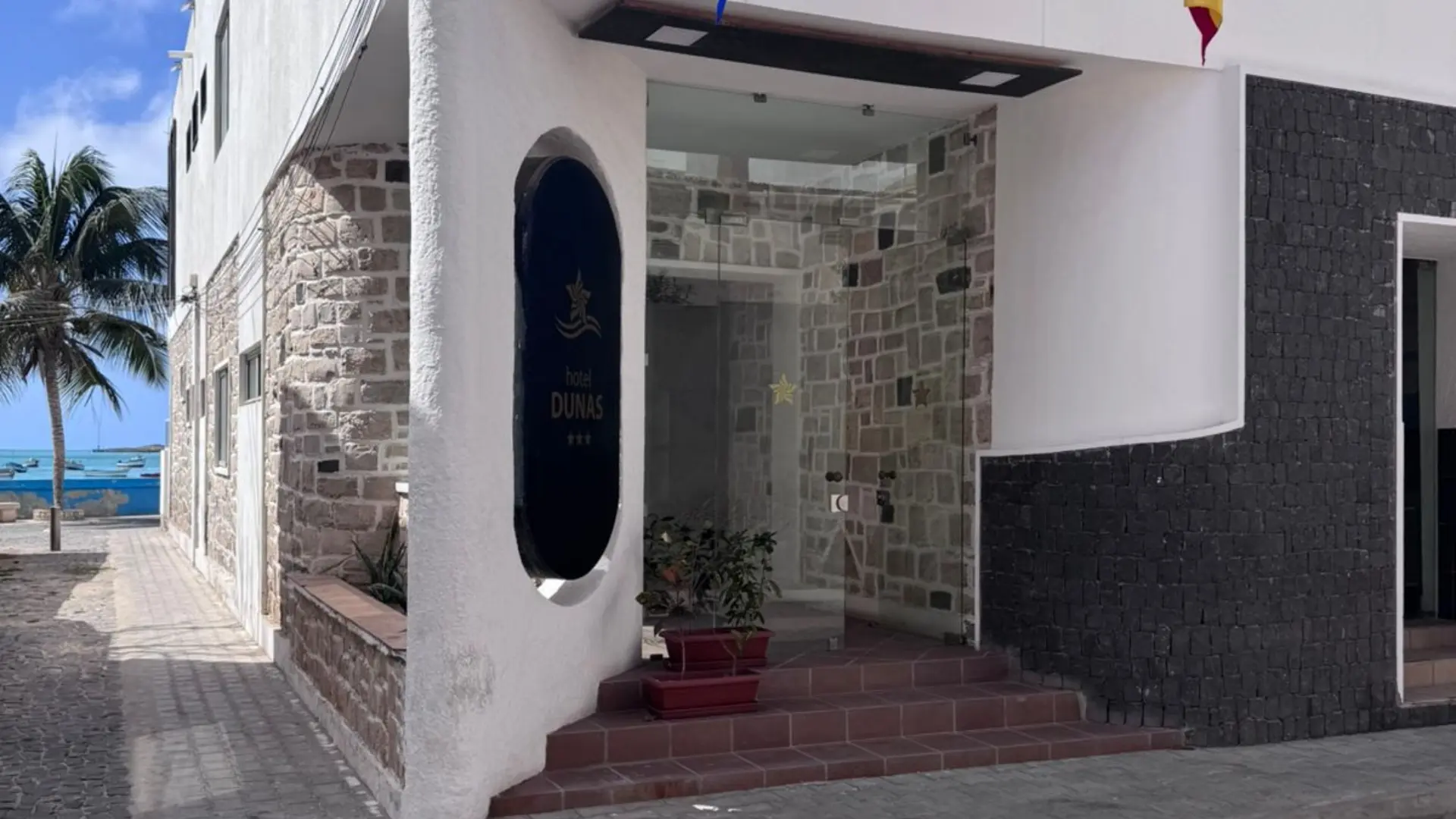 Entrada de hotel com fachada de pedra, plantas em vasos e rua de paralelep&iacute;pedos com a praia e uma palmeira ao fundo.
