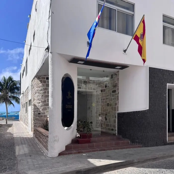 Entrada de hotel com fachada de pedra, plantas em vasos e rua de paralelep&iacute;pedos com a praia e uma palmeira ao fundo.