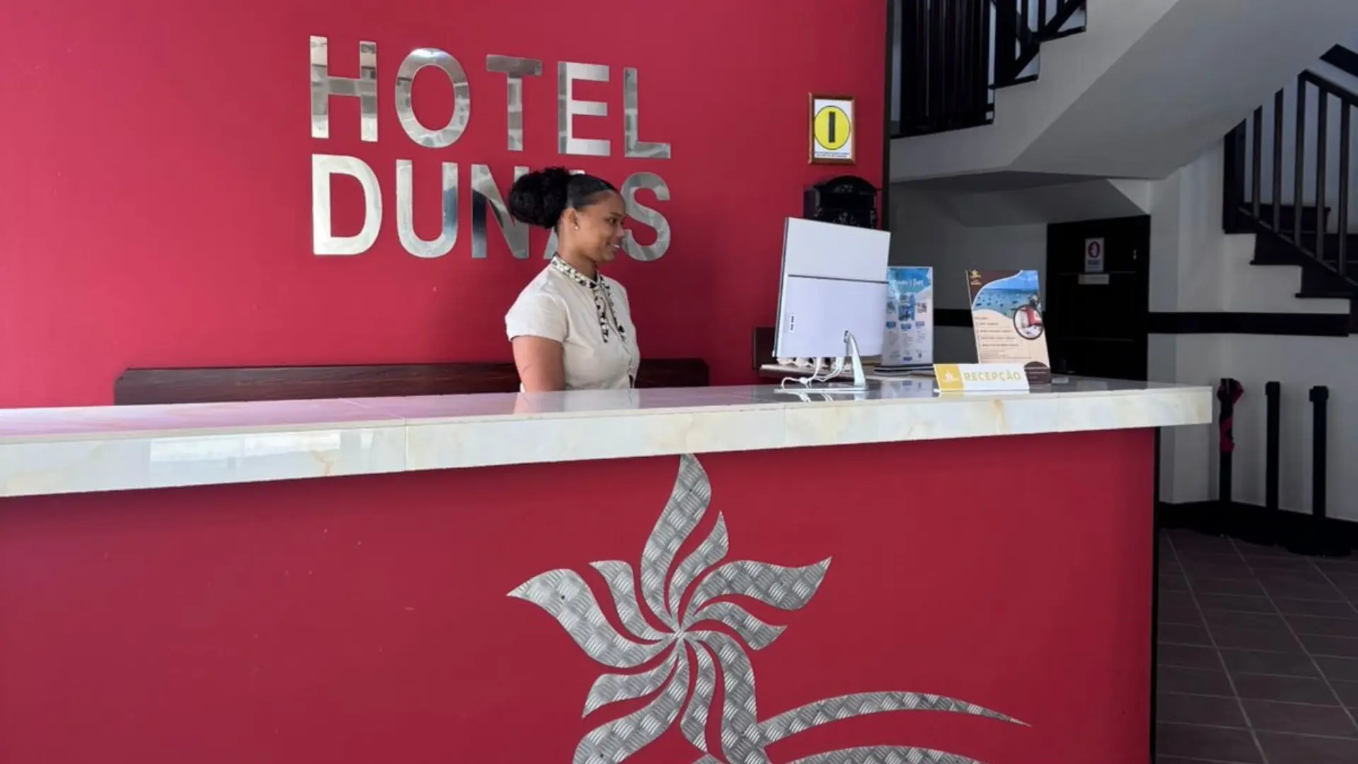 Recep&ccedil;&atilde;o do Hotel Dunas com atendente ao computador, fundo vermelho e logotipo na parede.