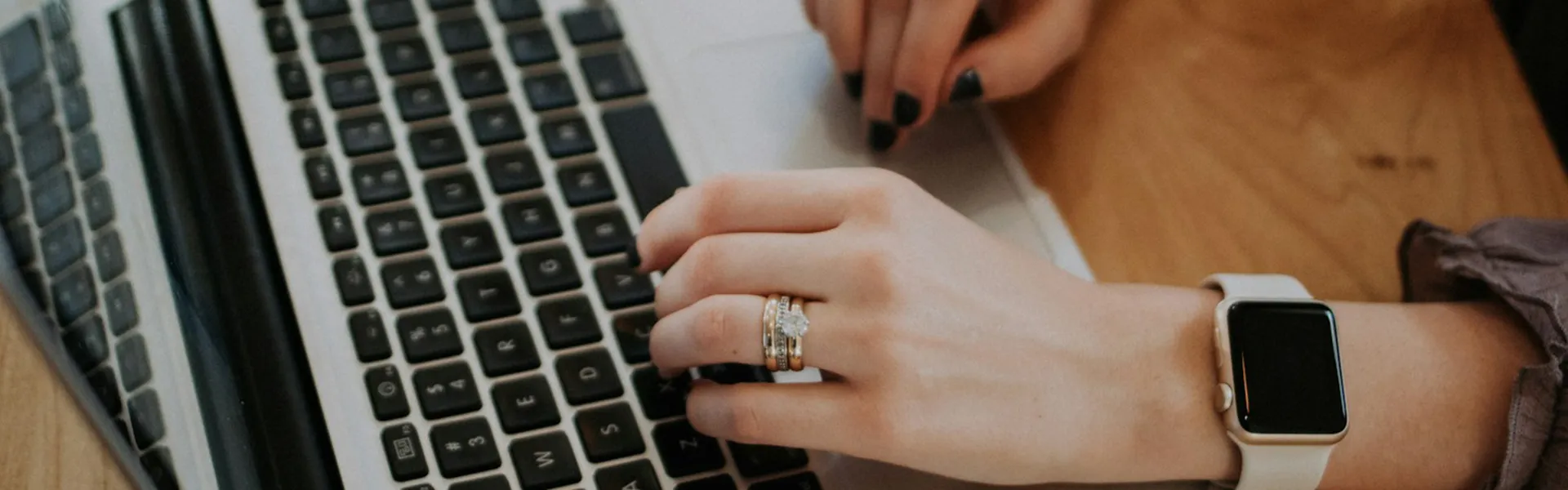 Manos escribiendo en un teclado de laptop, una con reloj y anillos.