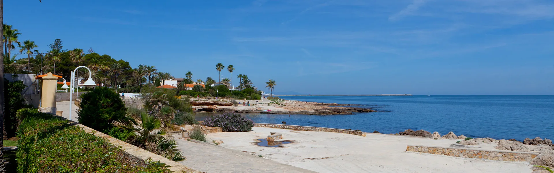 Costa con palmeras, casas y un camino junto al mar en un d&iacute;a soleado.