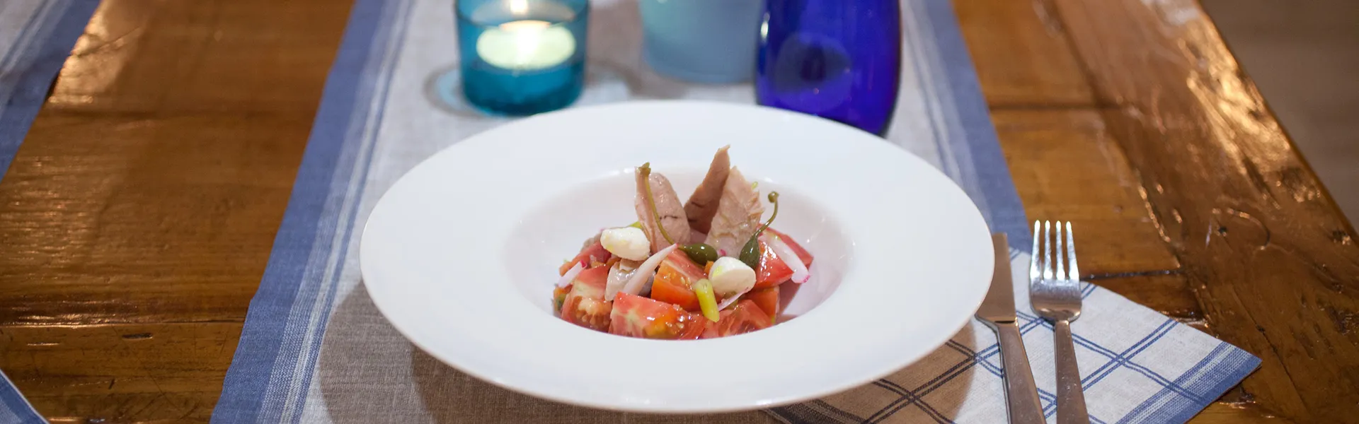 Plato de ensalada con at&uacute;n, sobre mantel a rayas junto a vela azul.