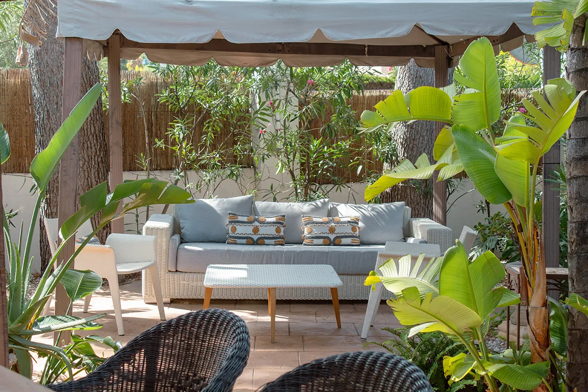 Patio con sofá, cojines decorativos, plantas y una mesa bajo un toldo.
