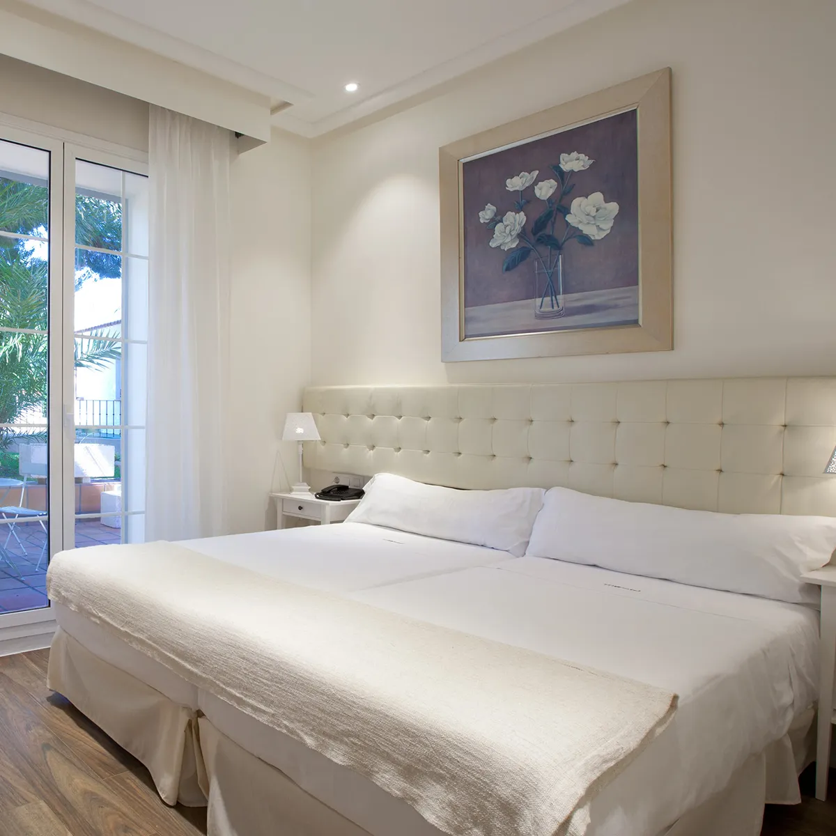 Habitaci&oacute;n de hotel con cama doble, ventana amplia y cuadro floral en la pared.