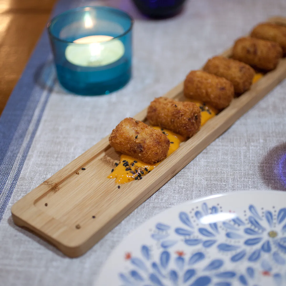 Croquetas en tabla de madera con salsa, junto a una vela azul y un plato decorado.