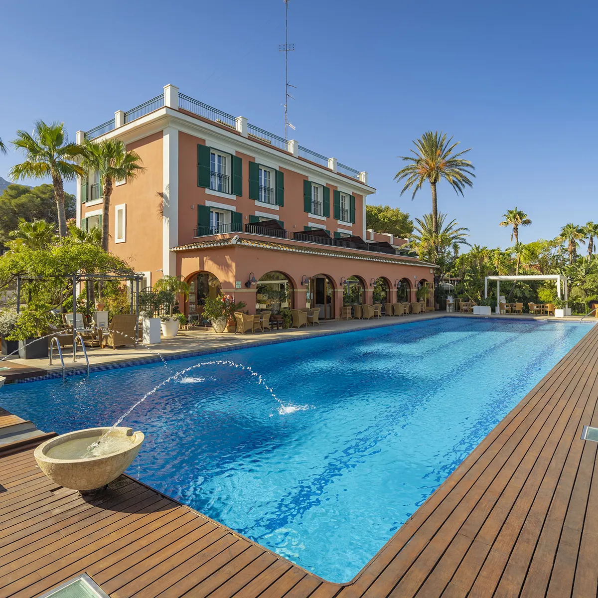 Mansi&oacute;n con piscina rodeada de palmeras y terraza con sillas al aire libre.
