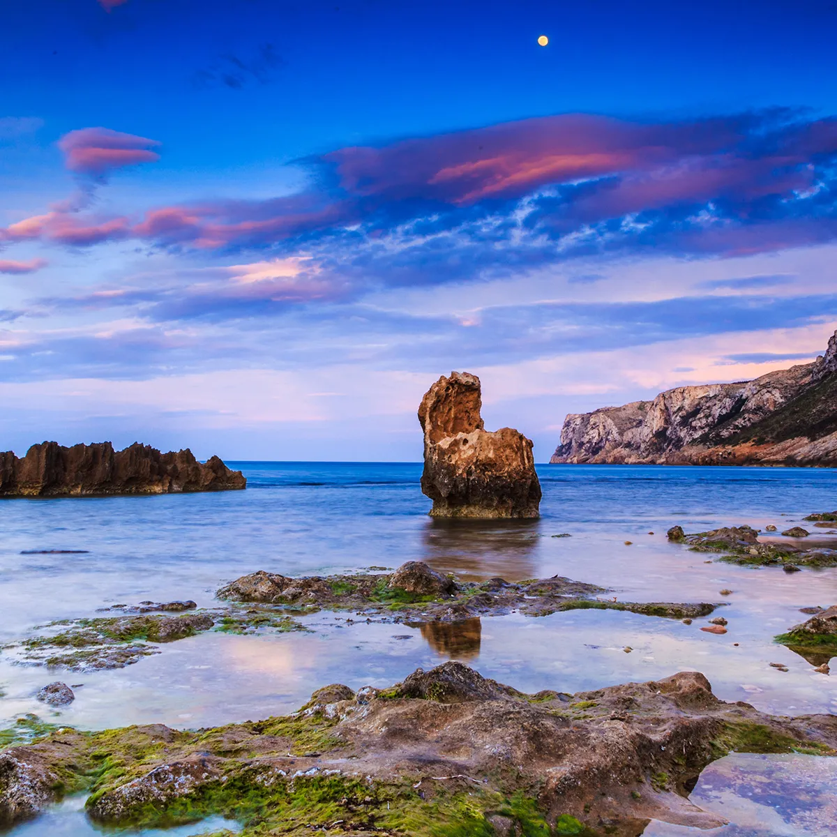 Playa rocosa al atardecer con cielo azul, nubes rosas y una luna visible.