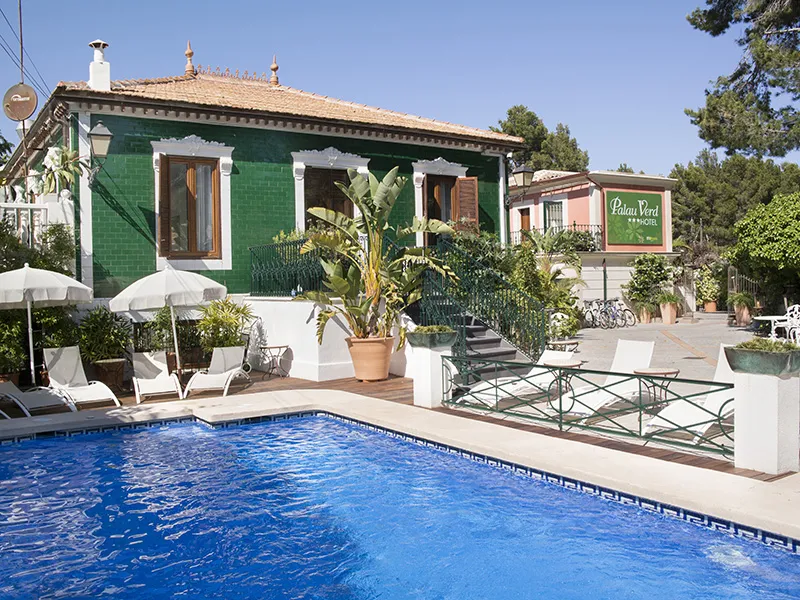 Casa verde con piscina y terraza soleada, rodeada de plantas y sombrillas.