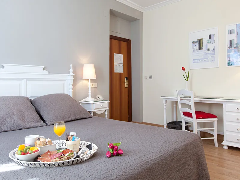 Habitaci&oacute;n de hotel con cama doble, desayuno y escritorio con silla blanca.