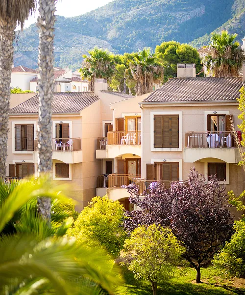 Edificios beige rodeados de palmeras y vegetaci&oacute;n, con monta&ntilde;as al fondo.