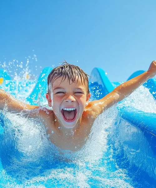 Ni&ntilde;o feliz desliz&aacute;ndose por un tobog&aacute;n de agua azul bajo un cielo soleado.