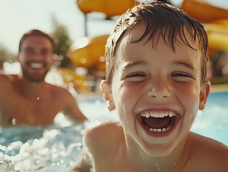 Un ni&ntilde;o sonr&iacute;e en una piscina con un adulto desenfocado detr&aacute;s.